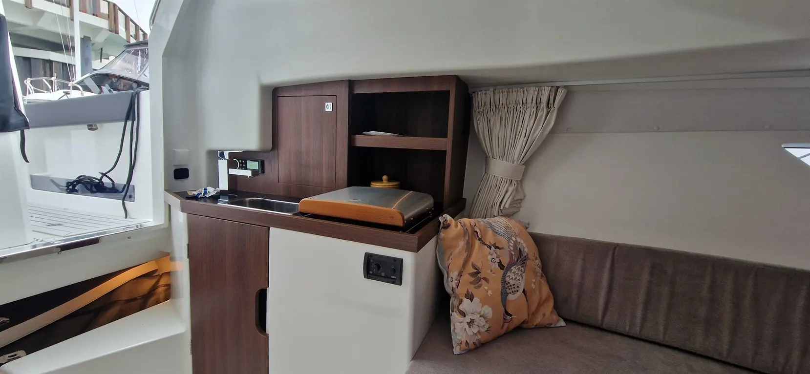 Modern yacht interior featuring a kitchenette with a sink, a wooden shelf, and decorative cushions on a grey sofa.